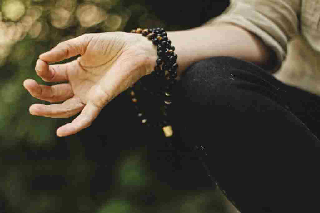 one hand in a yoga chin mudra placed on knee with a bracelet. man meditating along with a mudras and mantras to an an online yoga classes