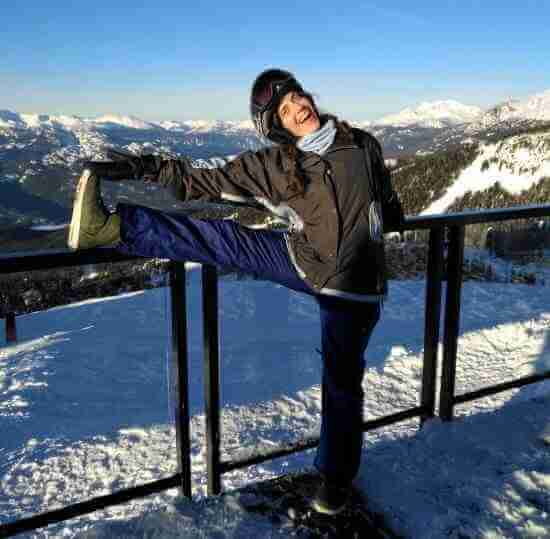 Heather Silberberg at the ski mountain, dressed in winter clothes. right leg is raised on to a hand rail. mountains in back ground