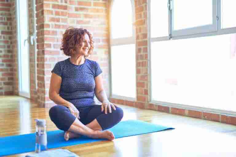 How Yoga Can Help With Pain Relief: Middle age woman wearing sportswear sitting on mat practicing yoga at home looking away to side with smile on face, natural expression. Laughing confident. Feeling less pain and less stress after practicing yoga. Migraine relief. Practicing to a yin yoga class