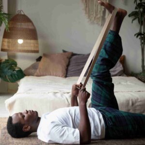 The Benefits of Using Yoga Props: Young african man working out at home , warming up using yogic belt, lying in yoga Supta Padangushthasana, One Leg Lift exercise, reclined variation of Extended Hand to Big Toe pose. a yoga strap for posture