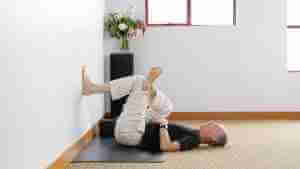 Bernie Clark laying down with one foot on the wall demonstrating an assisted figure four pose for the online Yin Yoga Teacher Training and classes