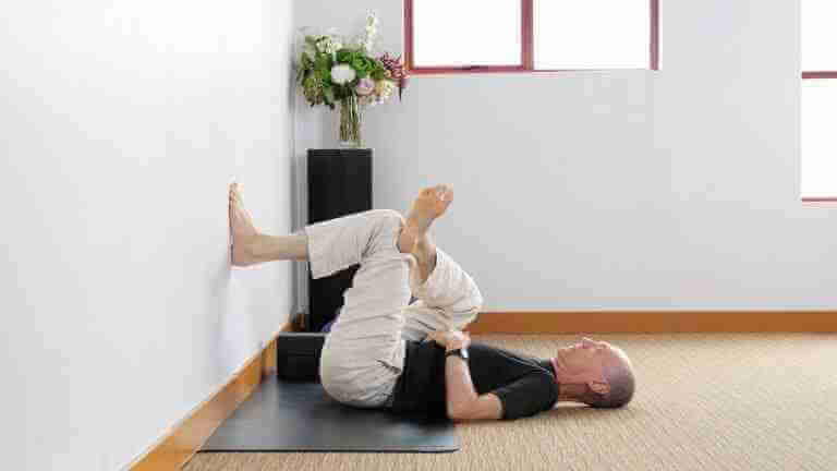 Bernie Clark laying down with one foot on the wall demonstrating an assisted figure four pose for the online Yin Yoga Teacher Training and online yin yoga classes. Yin Yoga Certification E-RYT