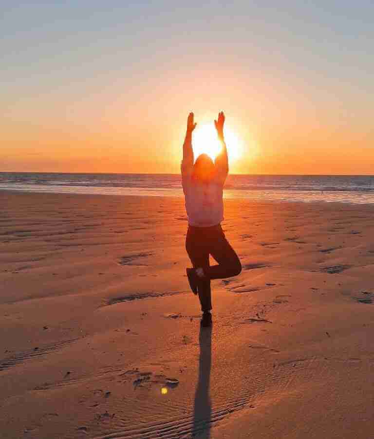 Anna Sierkova doing a yoga pose on a beach. the yoga pose is called tree pose. sunset in the background.
