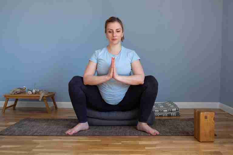 Pregnant woman practicing yoga squat pose Malasana sitting on yoga blocks and props. Using props to support her sacrum so she can do the pose and get the Yoga squat pose benefits