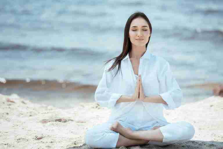 woman siting on a beach. legs crossed hands in prayer. Anjali Mudras and mantras