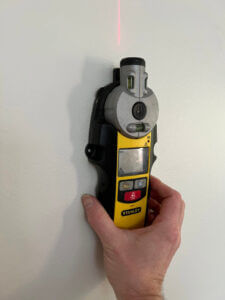wood stud finder being held to a white wall. looking for a wood stud to mount DIY Yoga Mat Storage