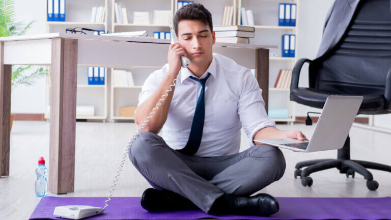 man sitting on a yoga mat on the floor of an office holding a phone. taking advantage of benefits of yoga in the workplace