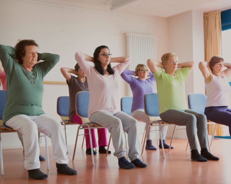 Mixed-ability group practicing chair yoga in a bright studio with a teacher offering gentle verbal cue.