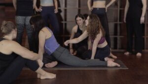 Yoga teacher guiding a student through embodied alignment cues while other students observe during teacher training