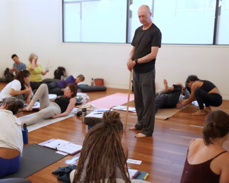 Bernie Clark Yoga Teacher walking slowly through the room or standing still, watching students with a calm, neutral expression