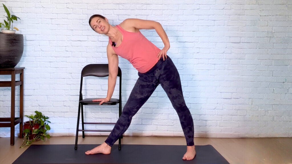 Christina Raskin in standing chair-supported Triangle Pose with her lower hand on a black chair seat.
