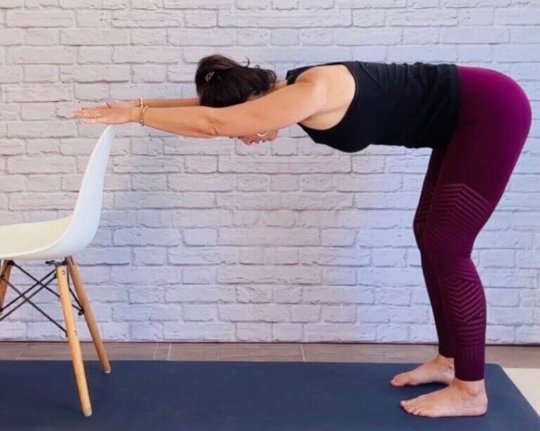 Christina Raskin doing standing chair yoga Downward Dog with both hands on a white chair backrest.