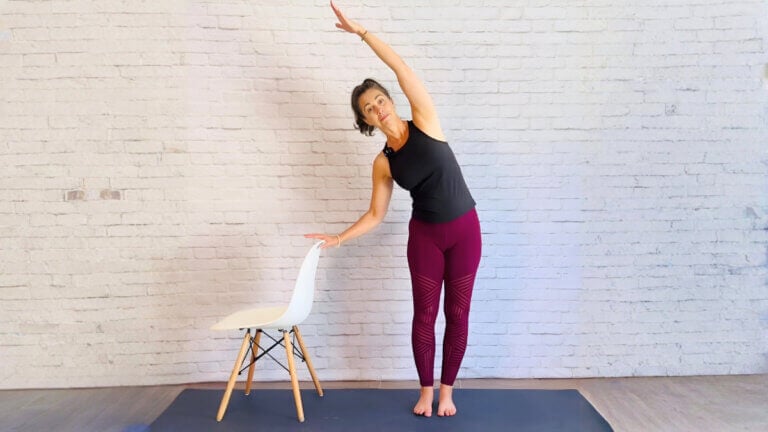 Christina Raskin doing a standing side bend beside a white chair with one hand on its backrest.